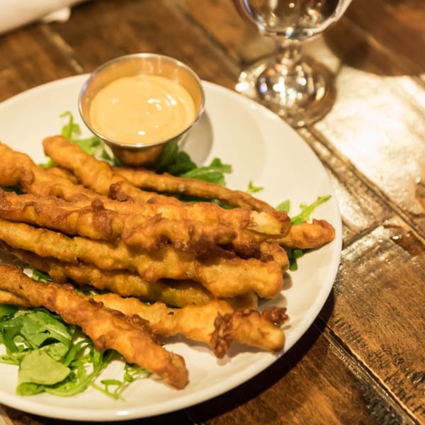 Crispy fried green bean fries with a side of dipping sauce on a white plate with arugula.