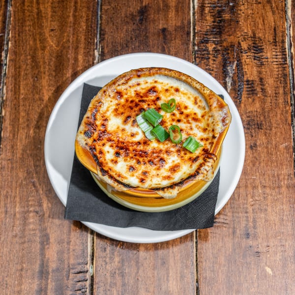 French onion soup in a brown crock with melted, bubbly cheese and green onions on a wooden table.