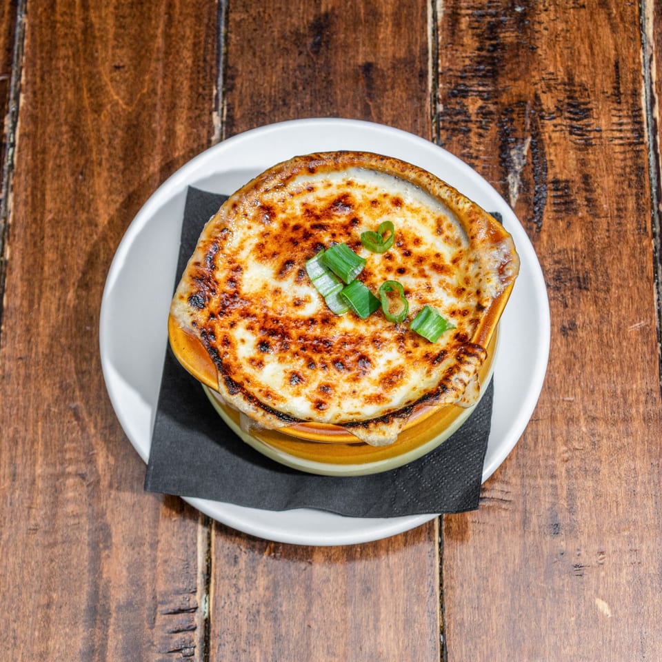 French onion soup in a brown crock with melted, bubbly cheese and green onions on a wooden table.