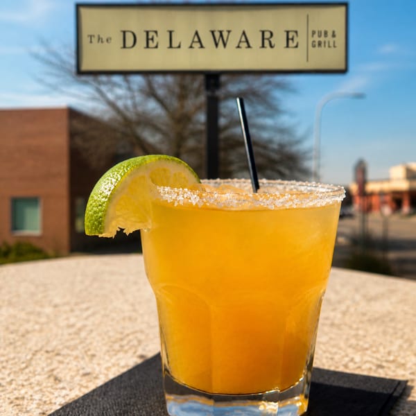 A vibrant orange margarita with a salted rim and lime wedge sits on a table outside The Delaware Pub & Grill.